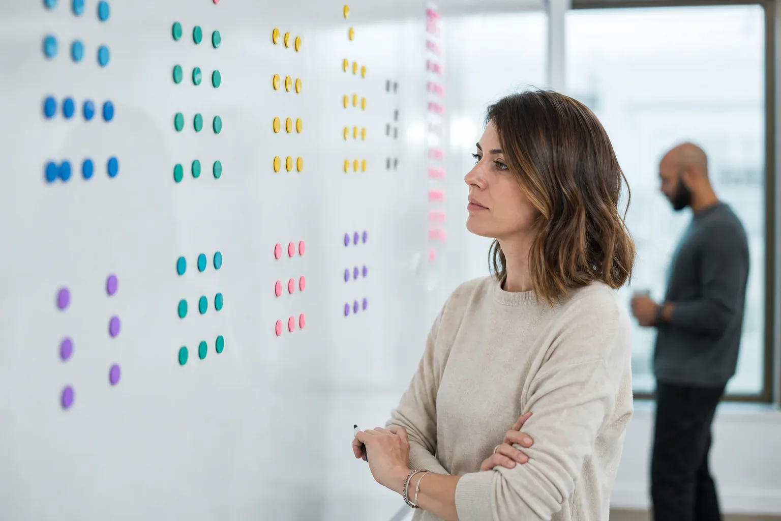 A large wall-mounted planning board with colour-coded magnetic markers arranged in intake-stage clusters, a digital marketing strategist in soft side light in the mid distance in sharp focus, a second figure blurred in the background, the board filling most of the frame.