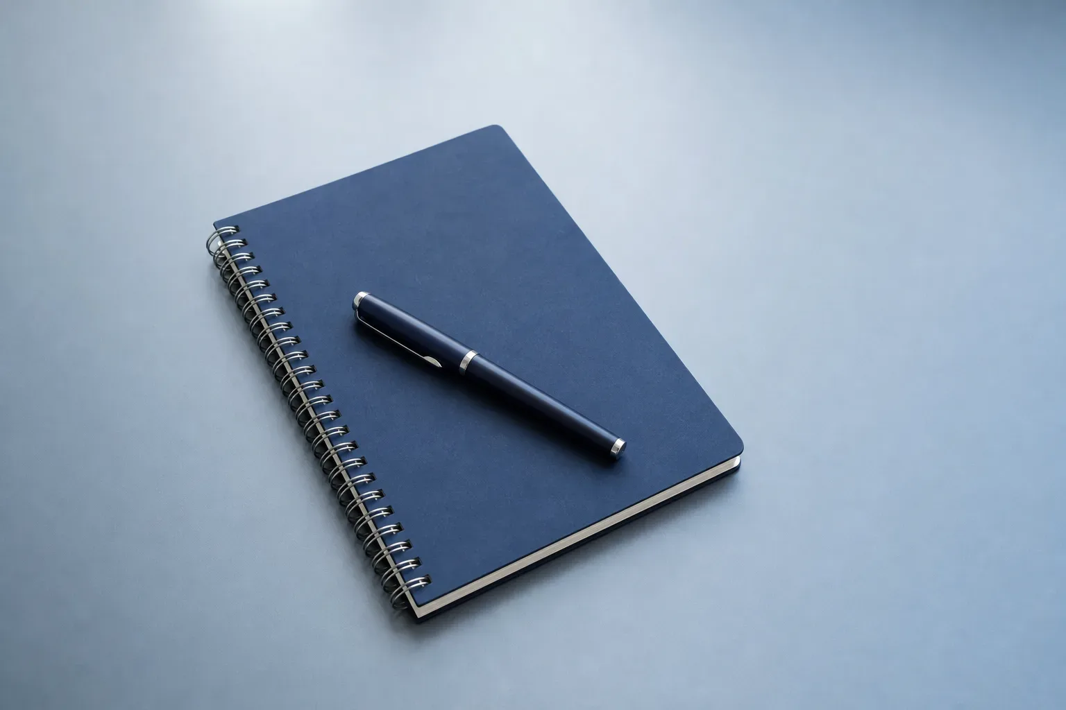 A closed spiral notebook with a capped pen across its cover on a clear desk, a wide view with cool ambient light from above, no person in frame, the notebook and pen the sole objects on the surface.