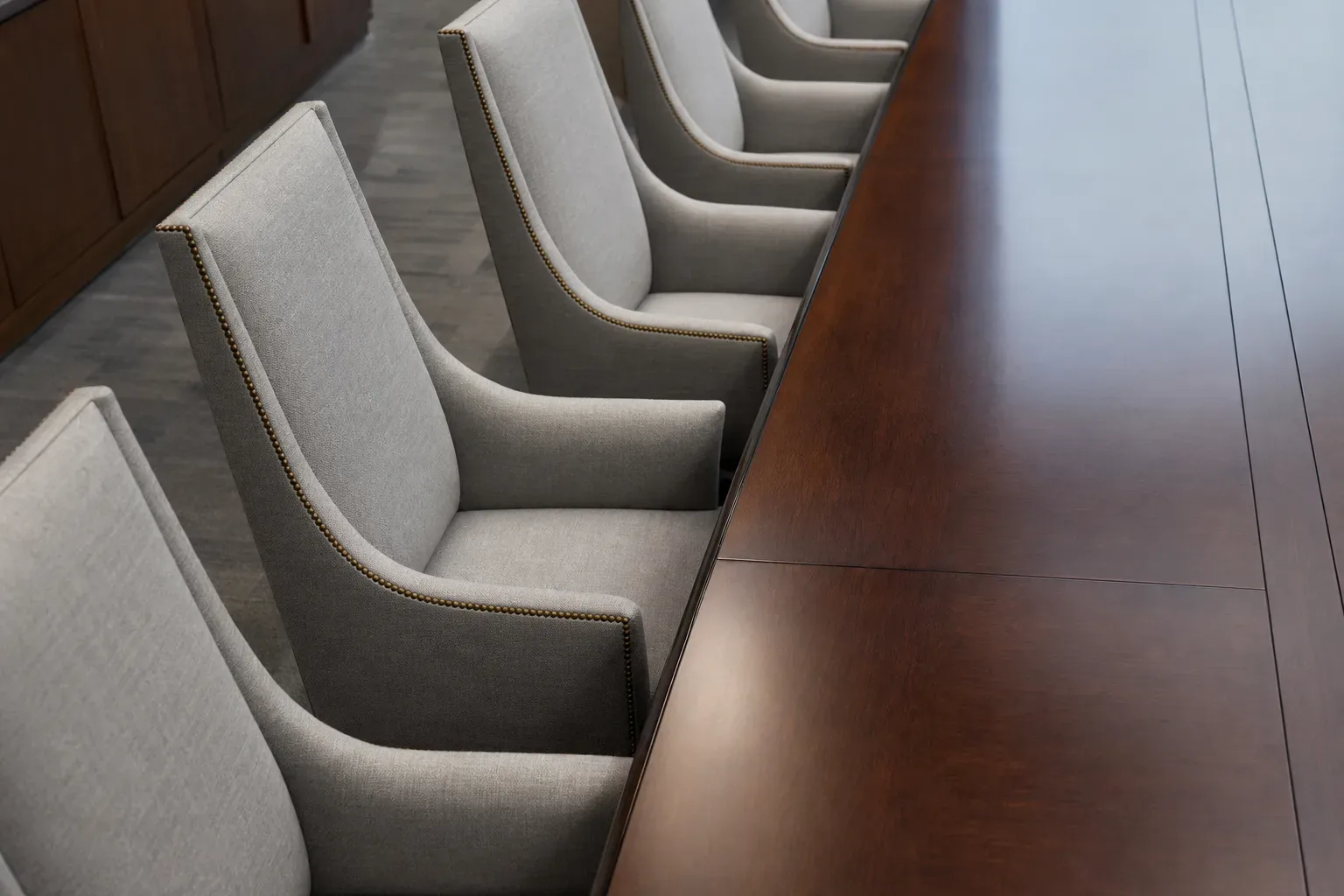 A row of high-backed upholstered chairs lines one side of a client meeting table, shot overhead in soft diffuse light, the evenly spaced chairs and polished table surface filling the frame, no person present.