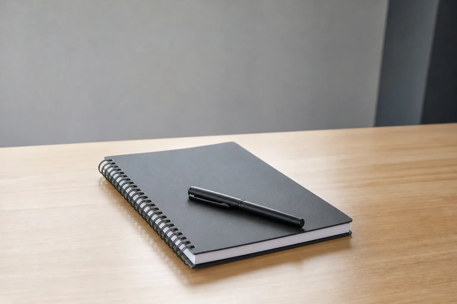A closed spiral notebook with a capped pen resting across its cover sits on a clear work surface, shot from a low angle looking upward toward the desk in calm even daylight, no person visible, the notebook the sole object on the surface.