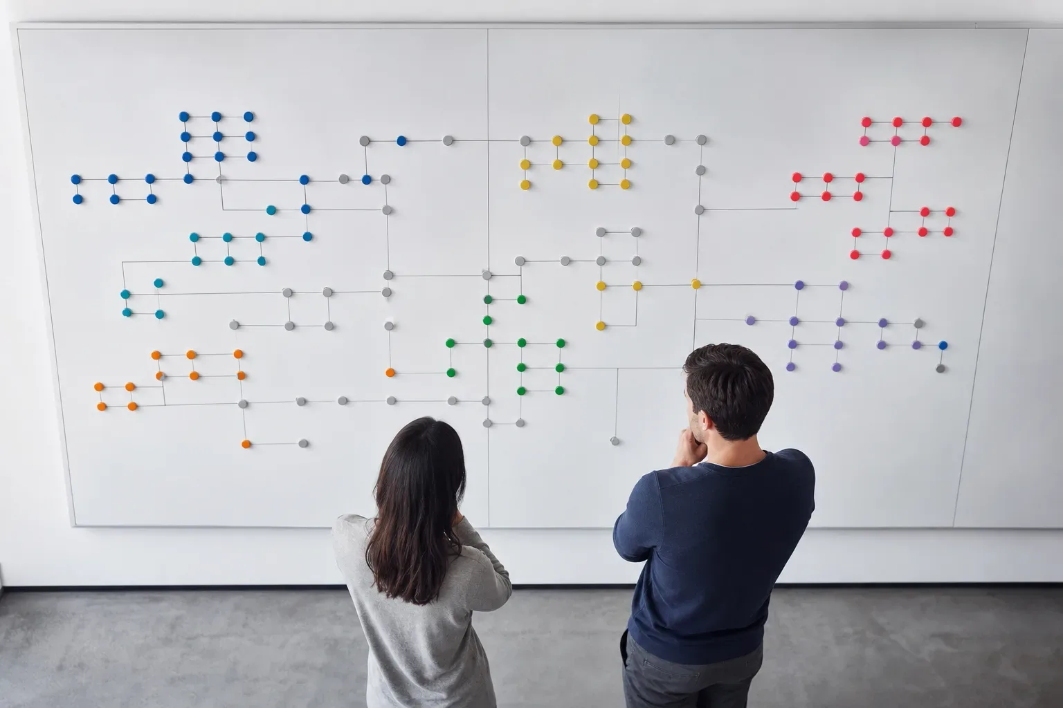 Two people stand with arms folded, studying a large wall-mounted board covered in colour-coded dot markers connected by lines