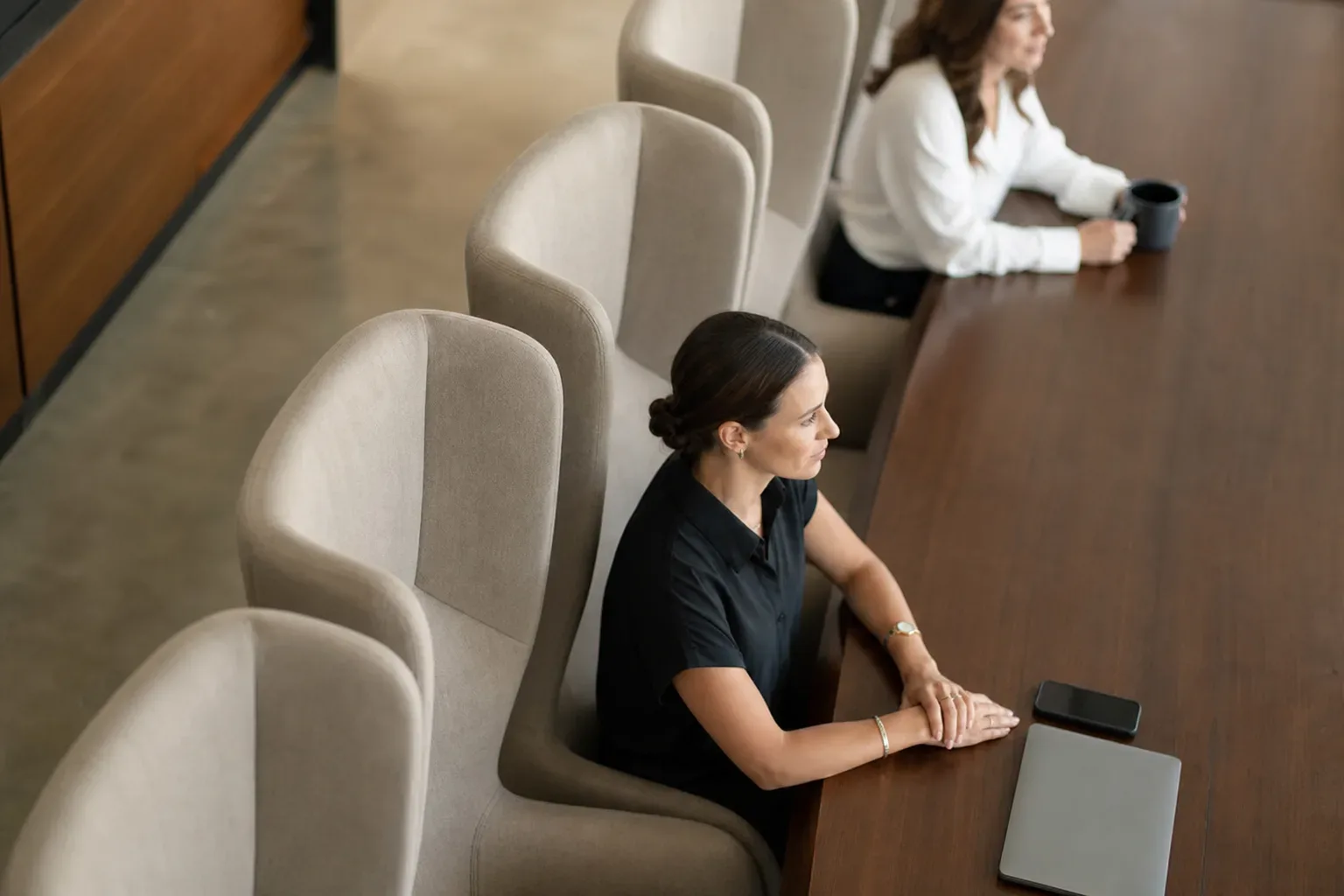 A row of high-backed upholstered chairs along one side of a client meeting table, seen from above in bright flat light, a digital marketing strategist's hands resting on the table surface at the near end, a second figure softly blurred further along.