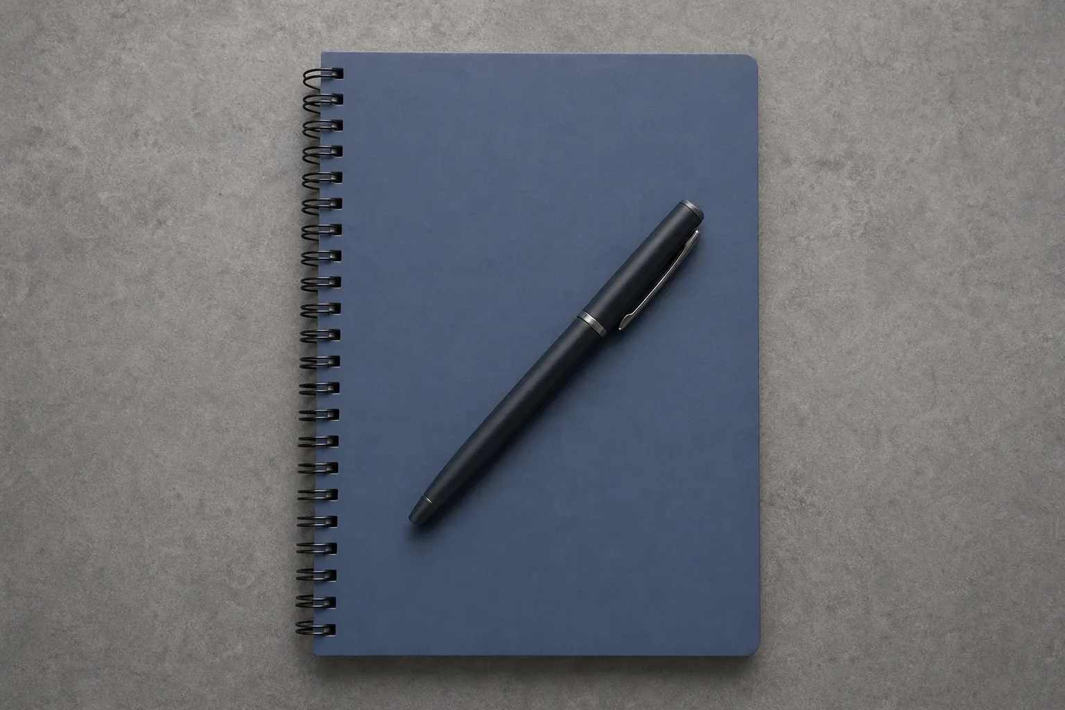 A closed spiral notebook with a capped pen across its cover on a clear work surface, shot overhead in close-up soft diffuse lighting, no person in frame, the notebook centred in the frame.