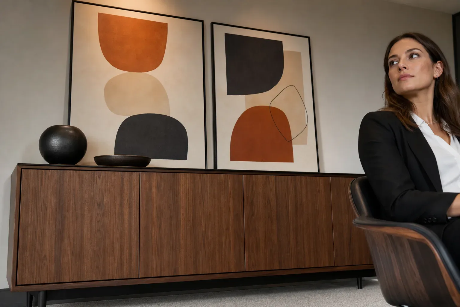 Timber credenza beneath framed abstract prints in a firm anteroom, shot from a low angle looking upward in calm even daylight, a digital marketing strategist partially visible at the side, the credenza and prints filling the upper frame.