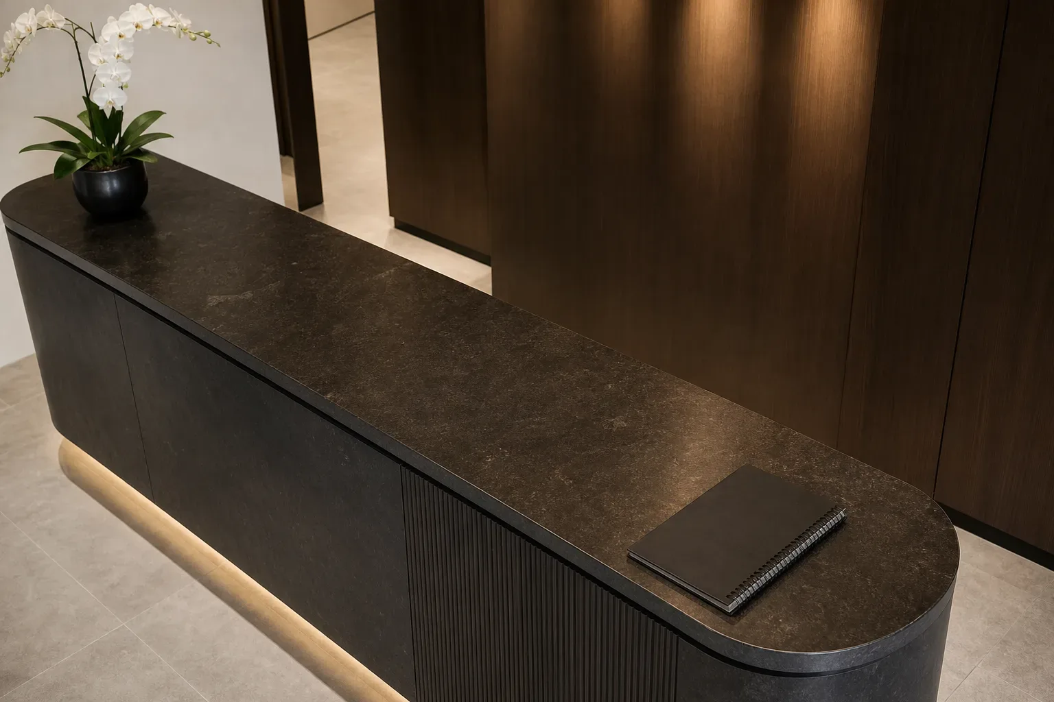A sleek law firm reception counter in dark stone, a single orchid at one end, seen from above in bright flat light, the stone surface bearing no objects other than a closed spiral notebook set at the near corner, nobody behind the counter.