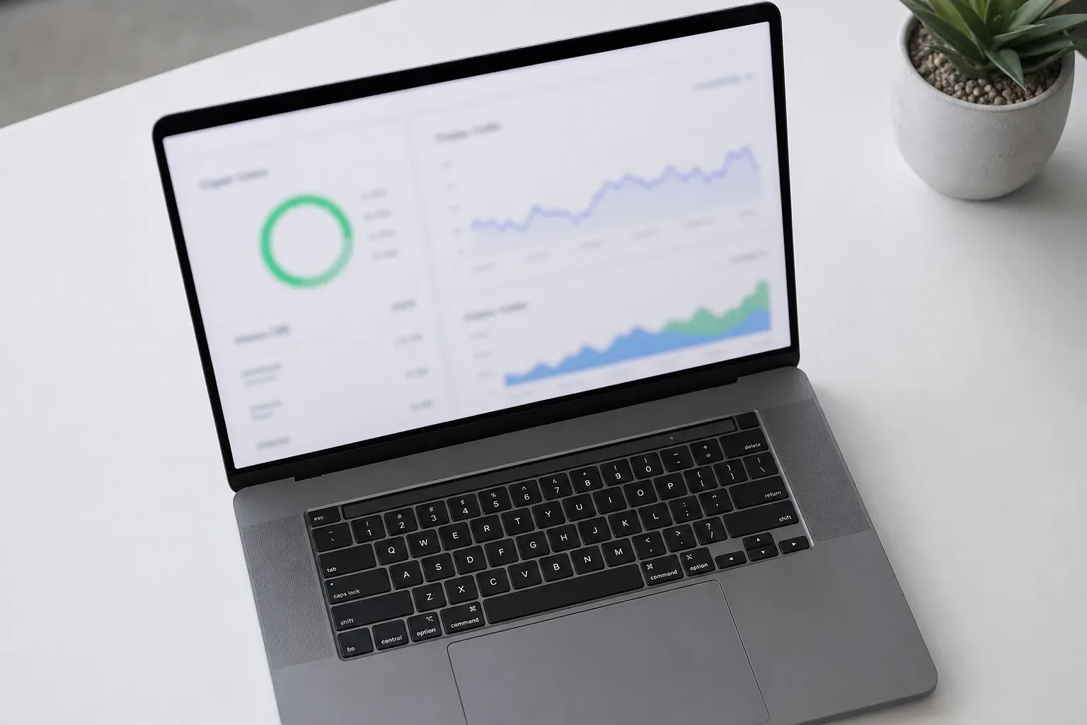 A laptop on a white surface displays a dashboard with a circular chart and line graphs, a small potted plant visible alongside